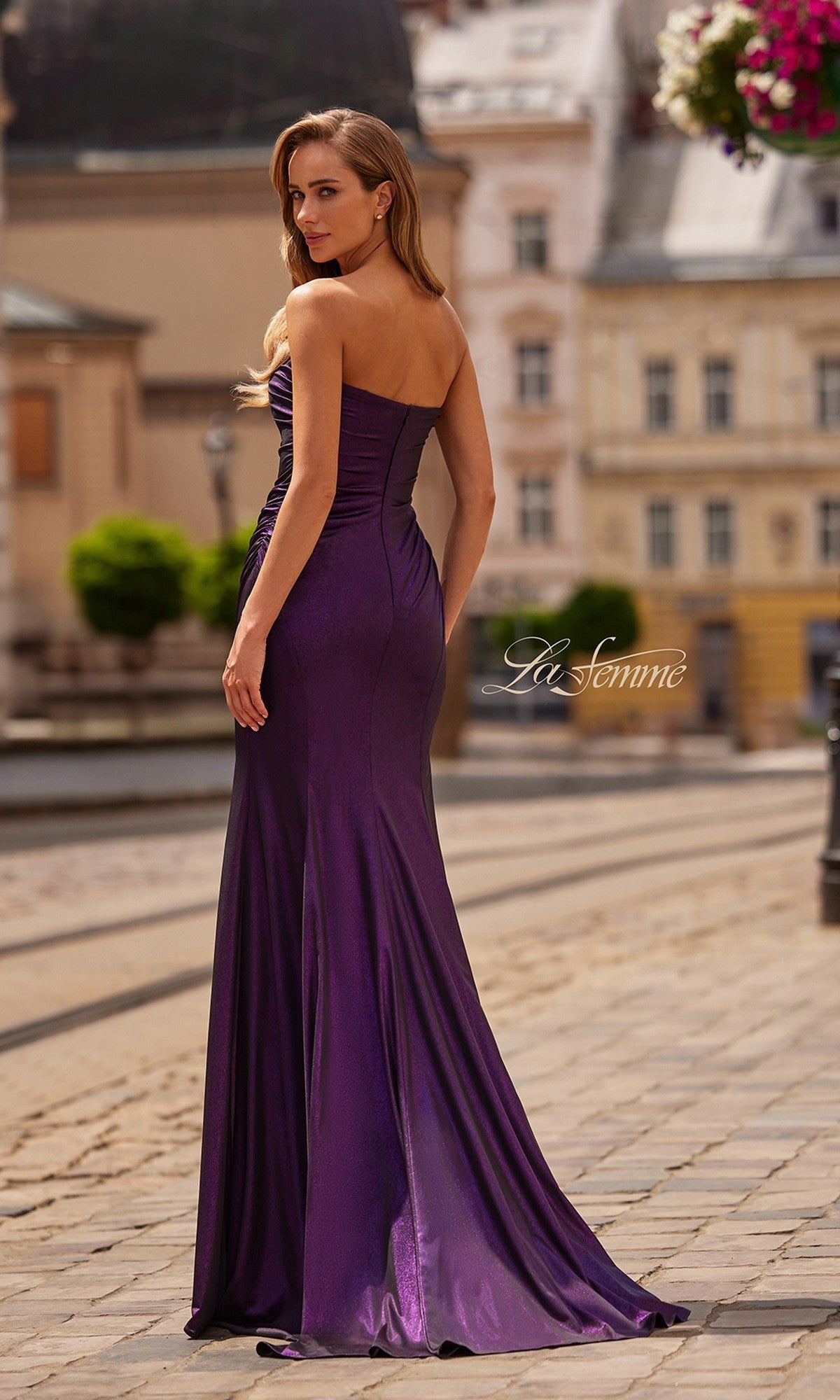 A woman in the La Femme 33424 long prom dress, featuring a strapless sweetheart neckline and floor-length purple design, stands outdoors on a cobblestone street, looking over her shoulder.